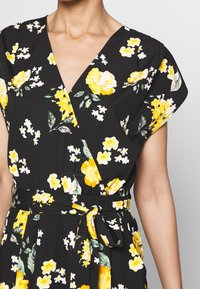Černé květinové šaty s žlutými květy a zelenými listy, s výstrhem do V, krátkými rukávy a detailem vázání v pase pro zvýraznění siluety.