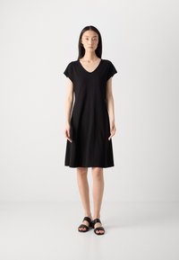 Black V-neck dress with short sleeves, loose fit, reaching mid-thigh, made of soft fabric. Paired with black heeled sandals.