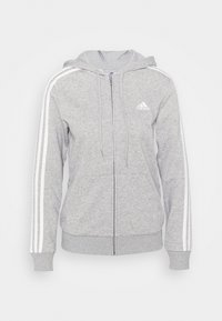 Šedá mikina s kapucí na zip vyrobená z měkké látky, má kapuci, přední kapsy a bílé pruhy na rukávech. Logo Adidas na hrudi.