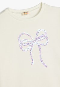Sudadera color crema con un diseño de lazo de lentejuelas multicolores y brillantes en el centro. Tejido suave con cuello y puños de canalé.