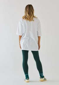 Biała oversize'owa koszulka, zielone legginsy i kolorowe skarpetki z napisem. Białe tło. Akcent na luźny krój i warstwowe tekstury.