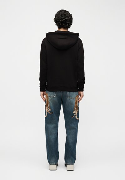 Personne aux cheveux bouclés portant un sweat à capuche noir et un jean bleu avec de grandes broderies de pieuvres sur les poches arrière, debout de dos.