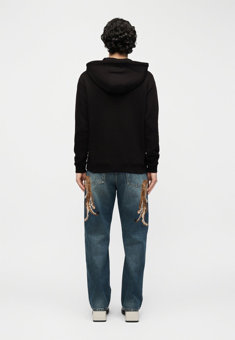 Persona con capelli ricci che indossa una felpa nera con cappuccio e jeans blu con grandi disegni di polpi ricamati sulle tasche posteriori, in piedi di spalle.