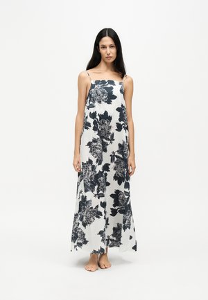 Woman with long dark hair wearing a white maxi dress with black floral print and thin straps, standing barefoot on a white background.