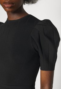 Haut noir ajusté en maille avec manches bouffantes texturées et détails côtelés porté par une personne à la peau foncée et aux cheveux naturels.
