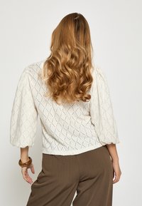 Biela blúzka s čipkou v tvare diamantového vzoru, trojštvrťovými nadýchanými rukávmi a mierne skráteným lemom, v kombinácii s hnedými širokými nohavicami.