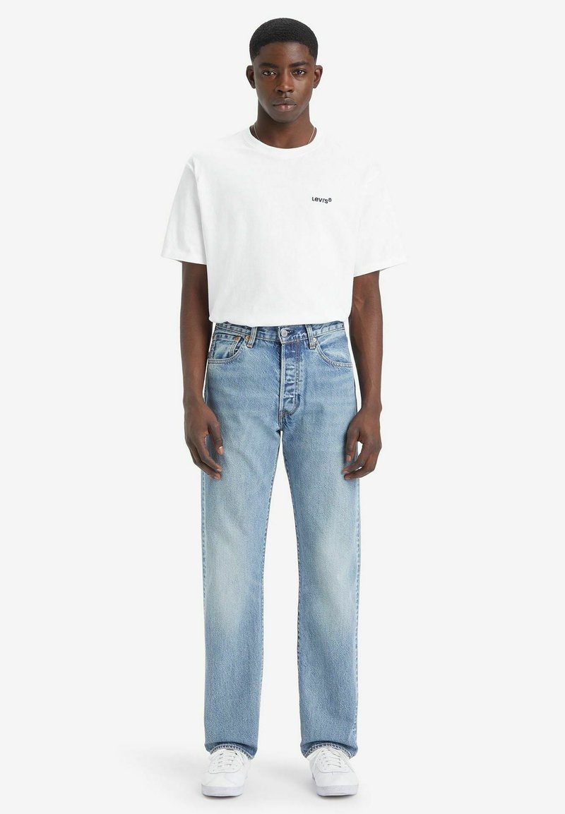 Witte katoenen t-shirt met zwart logo, gecombineerd met lichtblauwe rechte jeans. Model draagt witte sneakers en staat tegen een effen achtergrond.