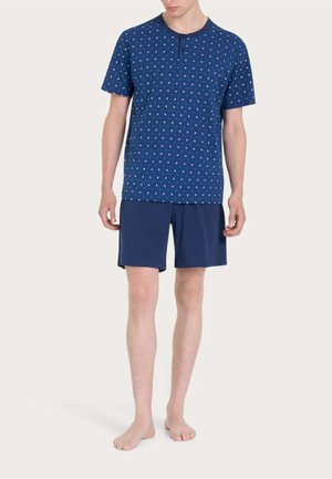 Uomo in piedi a piedi nudi che indossa una camicia blu a maniche corte con motivo e pantaloncini blu tinta unita su sfondo uniforme.