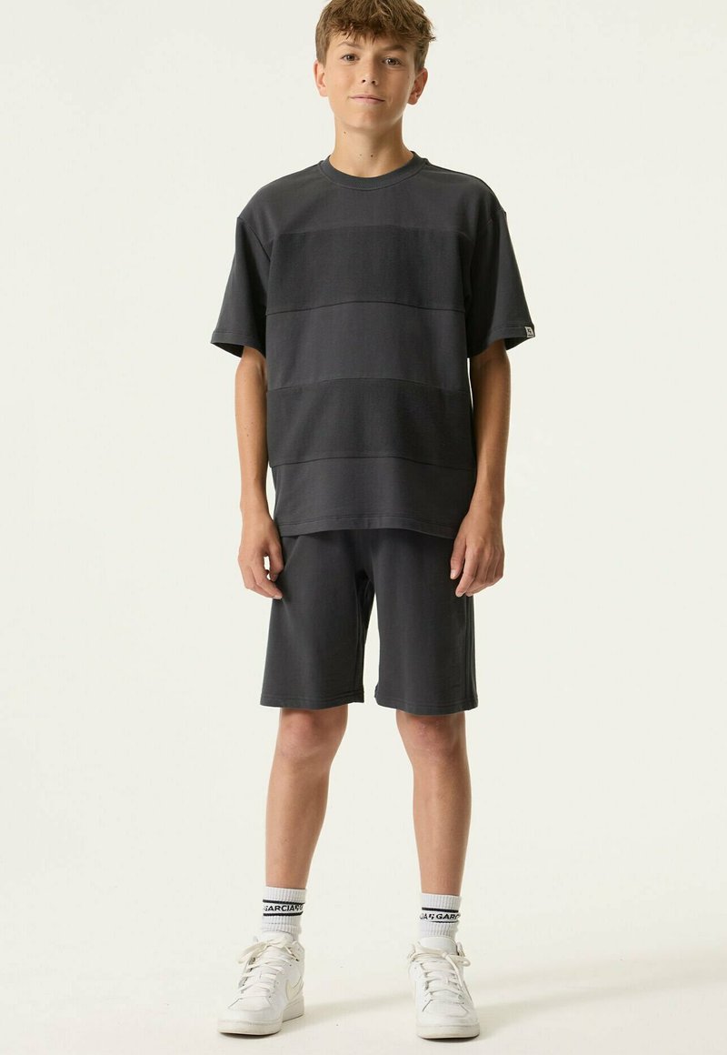 Junge steht und trägt ein dunkles, übergroßes T-Shirt sowie passende Shorts mit weißen Turnschuhen und markanten weißen Socken vor hellem Hintergrund.