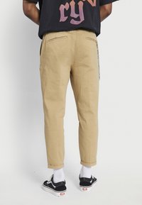 Khaki chinos with a tapered fit, featuring side pockets, cuffed hems, and a chain detail on the right side. Paired with black sneakers and white socks.