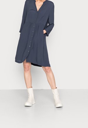 Femme portant une robe chemise bleu marine à manches longues et des bottines blanches, debout avec une main dans la poche sur un fond uni.