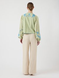 Kvinne med håret samlet bakover iført en lys grønn brodert bluse med blå design og beige vide bukser, stående vendt bort.