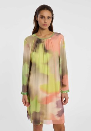 Frau trägt ein langärmeliges, knielanges Kleid mit abstrakten Pastellfarben in Grün, Pfirsich, Beige und Braun vor einem schlichten hellen Hintergrund.