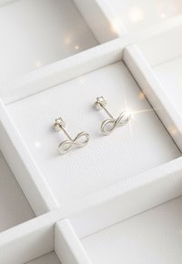 Orecchini a perno d'argento a forma di infinito con finitura lucida, esposti su uno sfondo bianco testurizzato. Presentano un design minimalista e un sistema di chiusura sicuro.