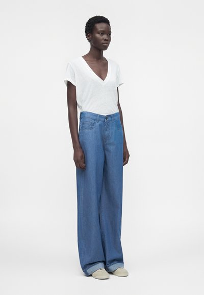 Γυναίκα όρθια με λευκό T-shirt με V λαιμόκοψη, ανοιχτό μπλε τζιν με φαρδιά πόδια και μπεζ αθλητικά παπούτσια σε απλό φόντο.