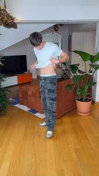 Jeune homme soulevant son t-shirt blanc pour montrer son abdomen, debout sur un sol en bois près d'un canapé marron et d'une grande plante verte dans le salon.