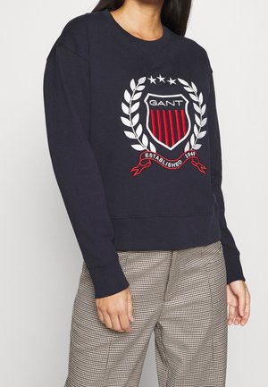 Persoon draagt een marineblauwe trui met rood-wit schildlogo en geruite broek, staand tegen een effen lichte muur.