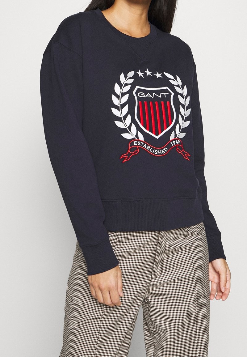 Personne portant un sweat-shirt bleu marine avec un logo écusson rouge et blanc et un pantalon à carreaux, debout contre un mur clair uni.