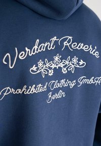 Hanorac din material albastru cu text brodat alb "Verdant Reverie" deasupra unui design floral și "Prohibited Clothing GmbH Berlin" dedesubt.