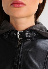 Black leather jacket with a gray fabric hood, featuring a silver zipper and snap closure, textured surface, and raised collar design.