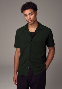 Camicia a maniche corte con bottoni in tessuto verde scuro, caratterizzata da un colletto scollato, una texture liscia e cinque bottoni neri sul davanti.