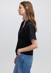 Top corto nero a maniche corte con scollatura a V, impreziosito da dettagli con borchie sulle spalle, abbinato a jeans azzurri chiari decorati con applicazioni.