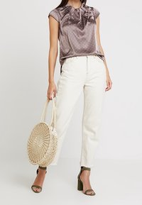 Korte mouwen, lila satijnen blouse met witte stippen, gecombineerd met off-white jeans en een ronde geweven tas. Olijfgroene sandalen met hakken zijn erbij te zien.