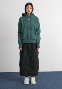 Groene hoodie met een voorzak, gecombineerd met een lange zwarte rok met ritsen. Witte sneakers maken de outfit af, tegen een effen achtergrond.