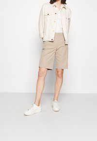 Beige Shorts aus Baumwoll-Twill, kombiniert mit einem weißen Hemd und einer hellbeigen Jeansjacke. Die Schuhe sind weiße Sneakers.