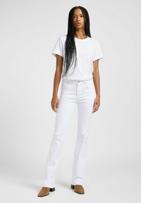 Camiseta blanca de algodón combinada con pantalones vaqueros acampanados de tiro alto en blanco. La modelo lleva zapatos de tac�ón de ante marrón y está de pie frente a un fondo claro.