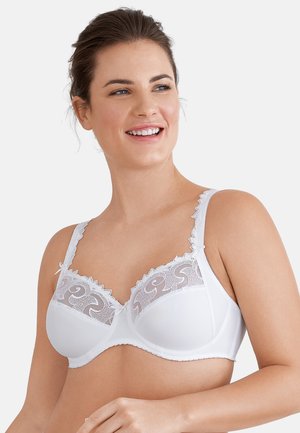 Kobieta uśmiechająca się, ubrana w biały biustonosz z fiszbinami z koronkowymi zdobieniami na górnej części miseczek i falbankowymi ramiączkami, na jednolitym jasnym tle.