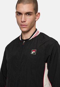 Svart bomberjacka med röd och vit randig krage, dragkedjestängning, broderad logotyp på bröstet och kontrasterande vita sidopaneler. Lättviktsmaterial.