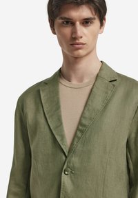 Helle olivgrüne Leinenblazer mit einem Einzelknopfverschluss, geschlitzten Revers und weicher Textur, getragen über einem beigen T-Shirt.