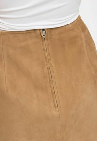 Gros plan d'une jupe en daim beige avec une fermeture éclair partiellement fermée à la taille, portée avec un haut blanc.