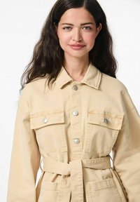 Veste en denim beige avec un col, boutons à l'avant, deux poches poitrine et une taille ceinturée. Texture douce et design classique.