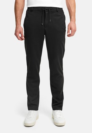 Man draagt strakke zwarte broek met trekkoord in de taille en witte sneakers, staand tegen een effen lichte achtergrond.