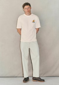 Hvid kortærmet t-shirt med farverigt logo, sammen med lys beige bukser og mørkebrune snørelædersko. Flad, struktureret baggrund.