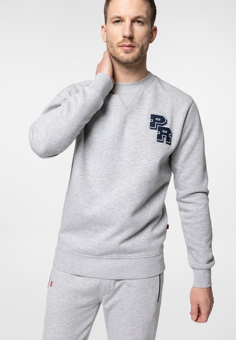 Szara bluza z miękkiego materiału z okrągłym dekoltem, ściągaczami przy rękawach i u dołu. Na piersi znajduje się granatowe logo.