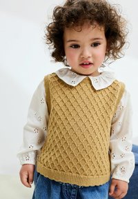 Gilet en tricot moutarde avec texture en losanges, porté par-dessus une blouse blanche avec des détails en dentelle et des manches bouffantes, associé à un jean.
