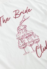 White fabric with a red outline drawing of a bottle pouring liquid into a pyramid of six glasses and red cursive text reading The Bride.