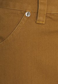 Tissu en denim marron avec une texture lisse, comprenant un bouton en métal portant le numéro 22 et des coutures visibles dans une couleur jaune contrastante.