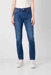 Persona che indossa jeans denim blu a taglio dritto, sneakers bianche e una maglietta bianca infilata, in piedi contro uno sfondo chiaro e uniforme.