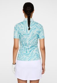 Jasnoniebieska koszulka golfowa z abstrakcyjnymi falistymi wzorami; krótki rękaw; gładka faktura; noszona z białą spódnicą i białą rękawiczką golfową.