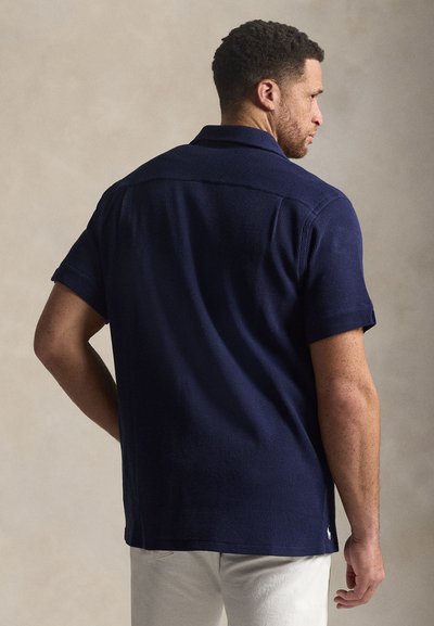 Μπλε σκούρο κοντομάνικο πουκάμισο από υφασμάτινη υφή με γιακά. Διαθέτει ίσια ρεβέρ και διακριτική λεπτομέρεια λογότυπου στο κάτω μέρος.