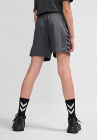 Grijze sportshorts met een gladde textuur en een elastische tailleband, voorzien van zwarte chevronpatronen aan de zijkanten; gecombineerd met zwarte sokken en schoenen.