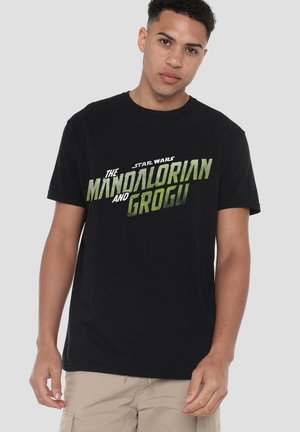 Junger Mann trägt ein schwarzes Star Wars T-Shirt mit grünem Text "The Mandalorian and Grogu" vor einem einfarbigen hellen Hintergrund.