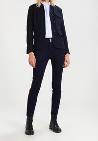 Giacca blu navy su misura con tasche frontali, indossata sopra una camicia azzurro chiaro. Pantaloni scuri aderenti con strisce laterali color bordeaux e stivali da combattimento neri.