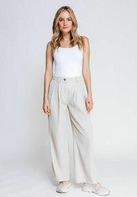 Beige weite Hosen aus strukturiertem Stoff, mit hoher Taille und einem einzigen Knopf vorne. Kombiniert mit einem weißen ärmellosen Top und Sneakers.