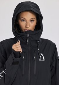 Giacca impermeabile nera con cappuccio, dotata di due tasche frontali con zip, cordini regolabili e dettagli del logo sul petto e sulla manica.