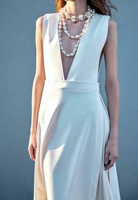 Femme portant une robe blanche sans manches avec un profond décolleté en V et plusieurs colliers de perles épaisses superposés sur un fond gris uni.
